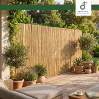 Natürliche Schilfmatte als Sicht- und Windschutz auf einer modernen Holzterrasse mit Sitzecke im Garten. Umgeben von Pflanzen schafft sie eine geschützte und gemütliche Terrasse. Oben rechts befindet sich das DE-COmmerce Logo.