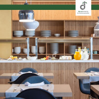Modern eingerichteter Restaurant-/ Bistrobereich mit hellem Bambus-Wandbelag, offenen Regalen mit Geschirr, Buffettheke mit Getränken und Speisen sowie gedeckten Holztischen und dunkelblauen Stühlen, oben rechts ist das DE-COmmerce Logo