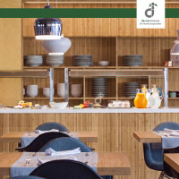 Modern eingerichteter Restaurant-/ Bistrobereich mit natürlichem Bambus-Wandbelag, offenen Regalen mit Geschirr, Buffettheke mit Getränken und Speisen sowie gedeckten Holztischen und dunkelblauen Stühlen, oben rechts ist das DE-COmmerce Logo.