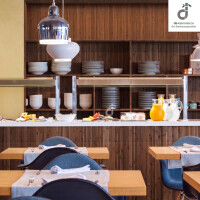 Modern eingerichteter Restaurant-/ Bistrobereich mit hellgrau-braunem Bambus-Wandbelag, offenen Regalen mit Geschirr, Buffettheke mit Getränken und Speisen sowie gedeckten Holztischen und dunkelblauen Stühlen, oben rechts ist das DE-COmmerce Logo.