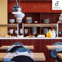 Modern eingerichteter Restaurant-/ Bistrobereich mit rotem Bambus-Wandbelag, offenen Regalen mit Geschirr, Buffettheke mit Getränken und Speisen sowie gedeckten Holztischen und dunkelblauen Stühlen, oben rechts ist das DE-COmmerce Logo.