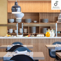 Modern eingerichteter Restaurant-/ Bistrobereich mit hellem Bambus-Wandbelag, offenen Regalen mit Geschirr, Buffettheke mit Getränken und Speisen sowie gedeckten Holztischen und dunkelblauen Stühlen, oben rechts ist das DE-COmmerce Logo.